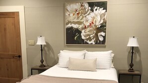 Cama bem arrumada com edredom branco e manta bege, com duas mesas de cabeceira com luminárias, uma porta de madeira e um quadro floral na parede.