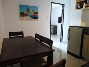 A dining area with a dark wooden table and chairs, a kitchen with a refrigerator and a microwave, and a painting of a boat on the wall. - The Uptown Place (Iloilo)