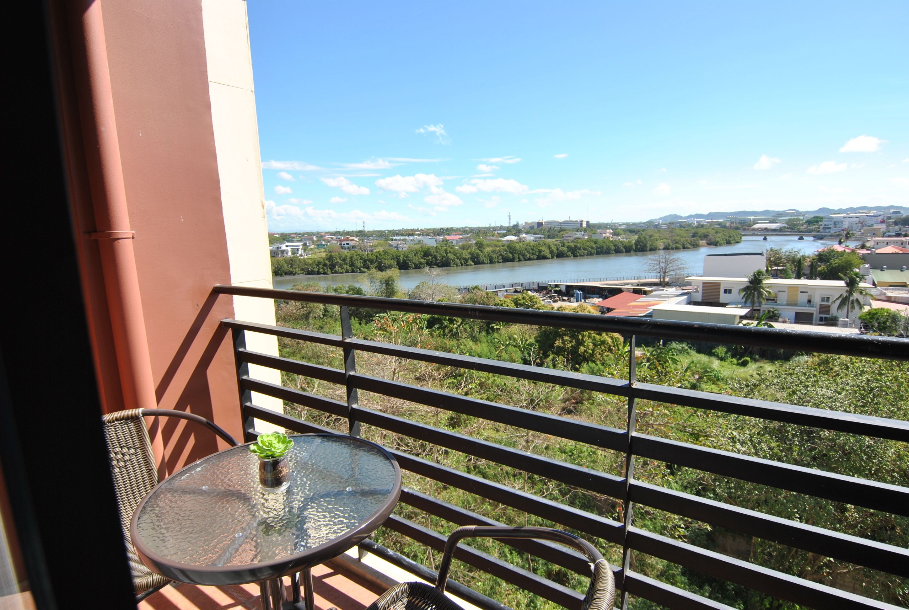 Varanda com mesa e cadeiras de vidro, com vista para o rio e a paisagem urbana.