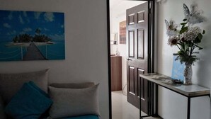 Une chambre à coucher avec un lit, un tableau, une console sur laquelle est posé un vase, et une vue sur la cuisine par une porte ouverte.