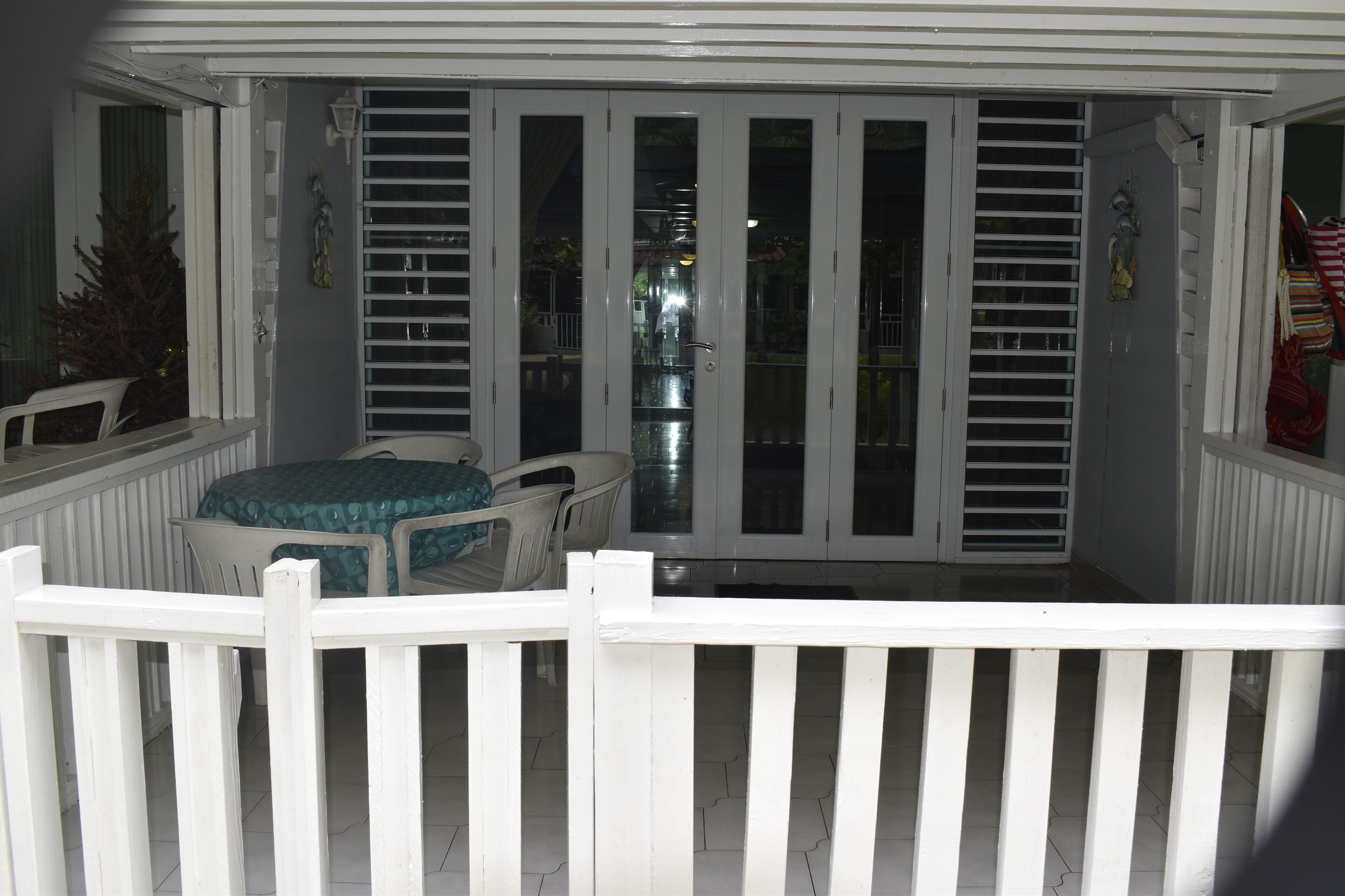 Une terrasse couverte avec une balustrade blanche, une table avec des chaises et une vue sur l’intérieur, visible à travers des portes vitrées.