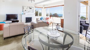 A modern living room with a glass dining table, beige sofa, and a TV. - BEACH BELLE - MORNINGTON (MORNINGTON)