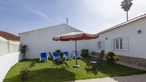 Eine Terrasse mit einem weißen Gebäude, gefliestem Boden und mehreren Liegestühlen unter einem großen Sonnenschirm.