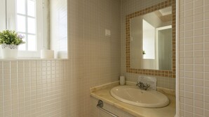 Ein Badezimmer mit weiß gefliesten Wänden, einem Waschbecken mit Spiegel, einem Fenster mit Pflanze und einer Haltestange.