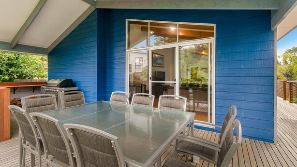 Une terrasse couverte avec une table et des chaises en verre, un barbecue et une vue sur l’intérieur de la maison grâce à une porte vitrée.