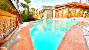 Una piscina con borde de piedra, rodeada por una casa de fachada de piedra y terraza de madera.