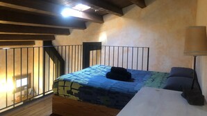 A bedroom with a sloped ceiling, a bed with a patterned bedspread, a black cat on the bed, a nightstand, a lamp, and a window with a view of the outside. - Rural apartment L'Alzina for 2 people (Tavertet)