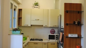 A compact kitchen with white cabinets, a microwave, a stove, and a refrigerator. There is a wooden door on the right and a small window on the left. - San Raffaele Città Studi Politecnico CIN IT015146C2F3B9XNN (Milano)