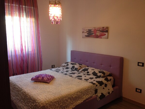 Une chambre avec une tête de lit violette, un lit recouvert d’une couverture à motifs de vaches, un oreiller rose et un tableau accroché au mur.