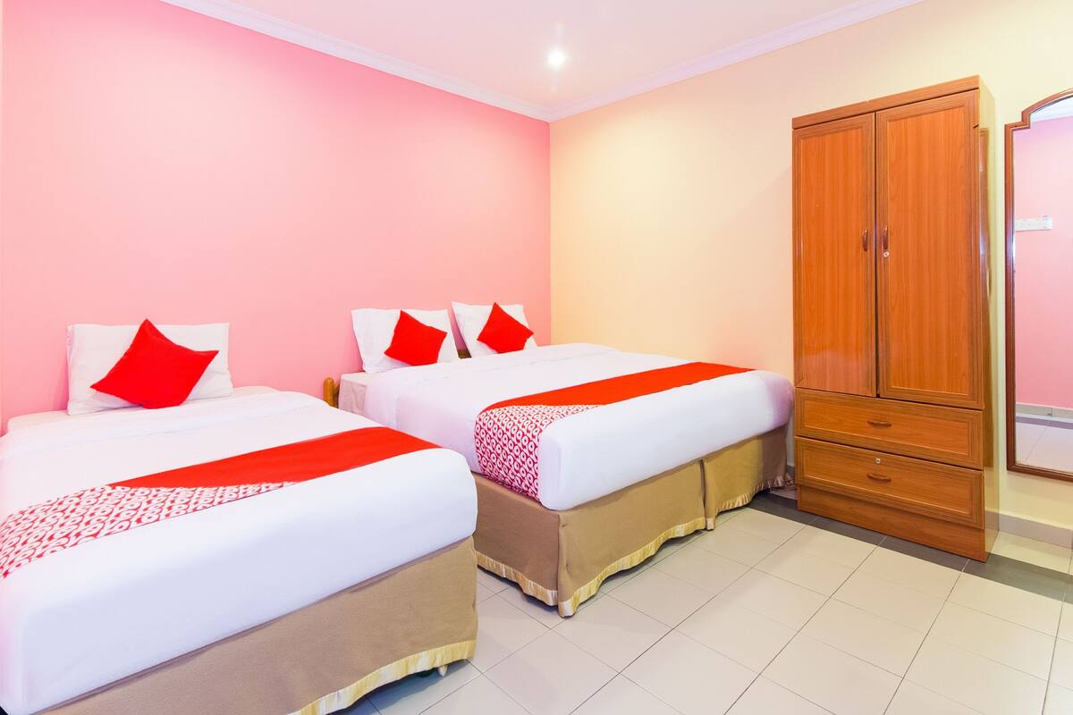 A hotel room with two beds, each with red pillows and white bedding, a wooden wardrobe, and a tiled floor.