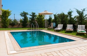 Piscina retangular com um deck revestido em azulejos, rodeada por espreguiçadeiras e um guarda-sol.