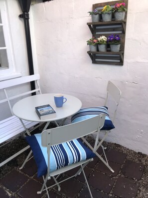Un petit espace extérieur aménagé avec une table et des chaises en métal blanc, une tasse bleue et un livre. À gauche, on aperçoit un banc blanc et, au mur, une étagère sur laquelle sont disposées des plantes en pot.