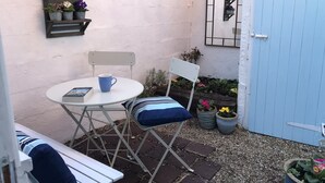 A small patio area with a white table and chair, a blue mug, and a book. There is a white door and a mirror on the wall. - Pretty 18th century cottage centrally situated  on Mermaid Street in Rye (Rye)