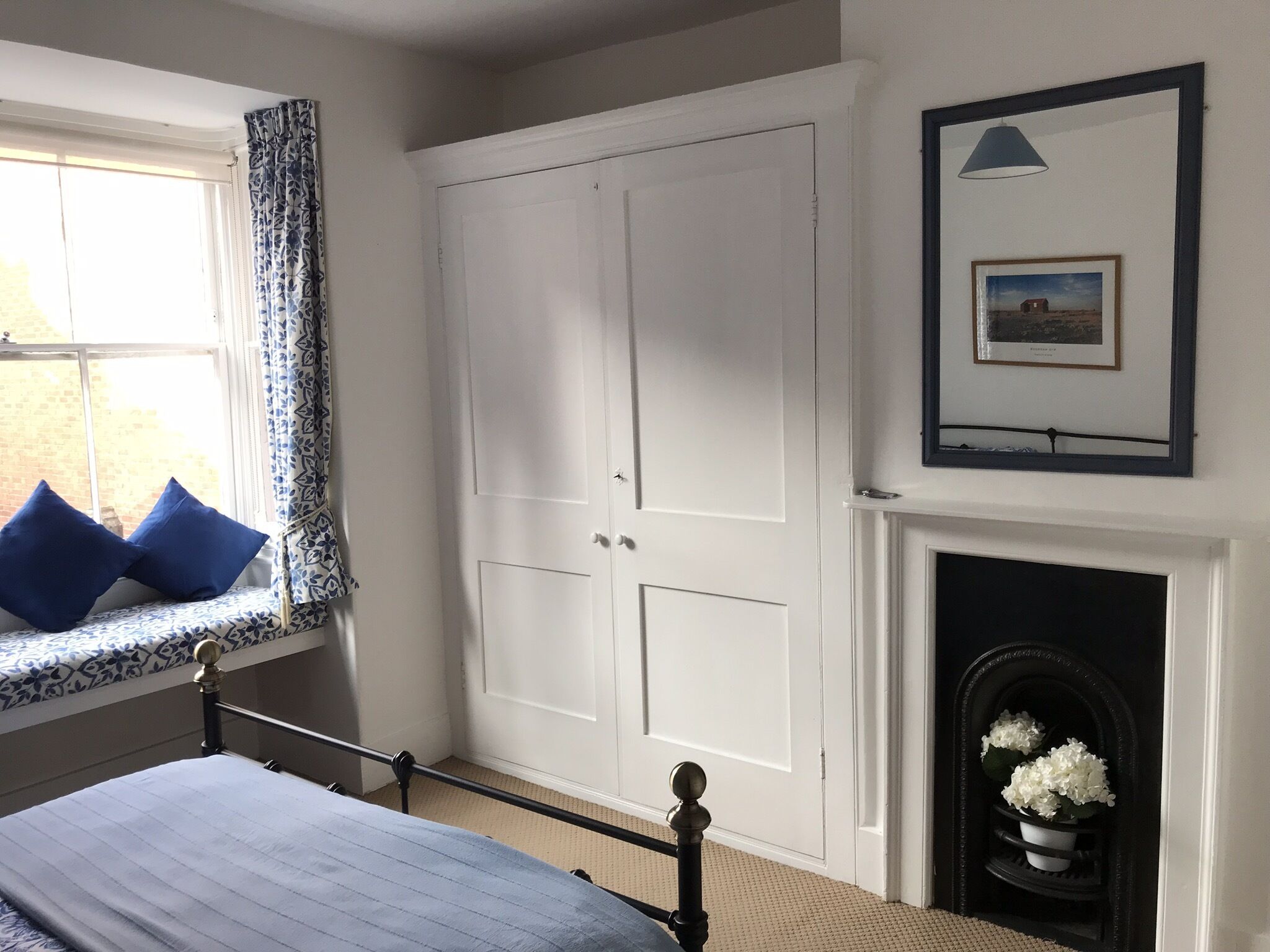 A bedroom with a bed, a window with blue curtains, a white wardrobe, a framed picture above a fireplace, and a small plant on the mantel.