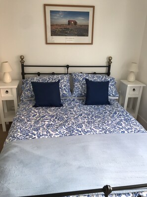 Un lit recouvert d’une couvre-lit à motifs floraux, agrémenté de deux oreillers, et flanqué de deux tables de chevet blanches sur lesquelles reposent des lampes, dans une pièce dont le mur est orné d’un tableau encadré.