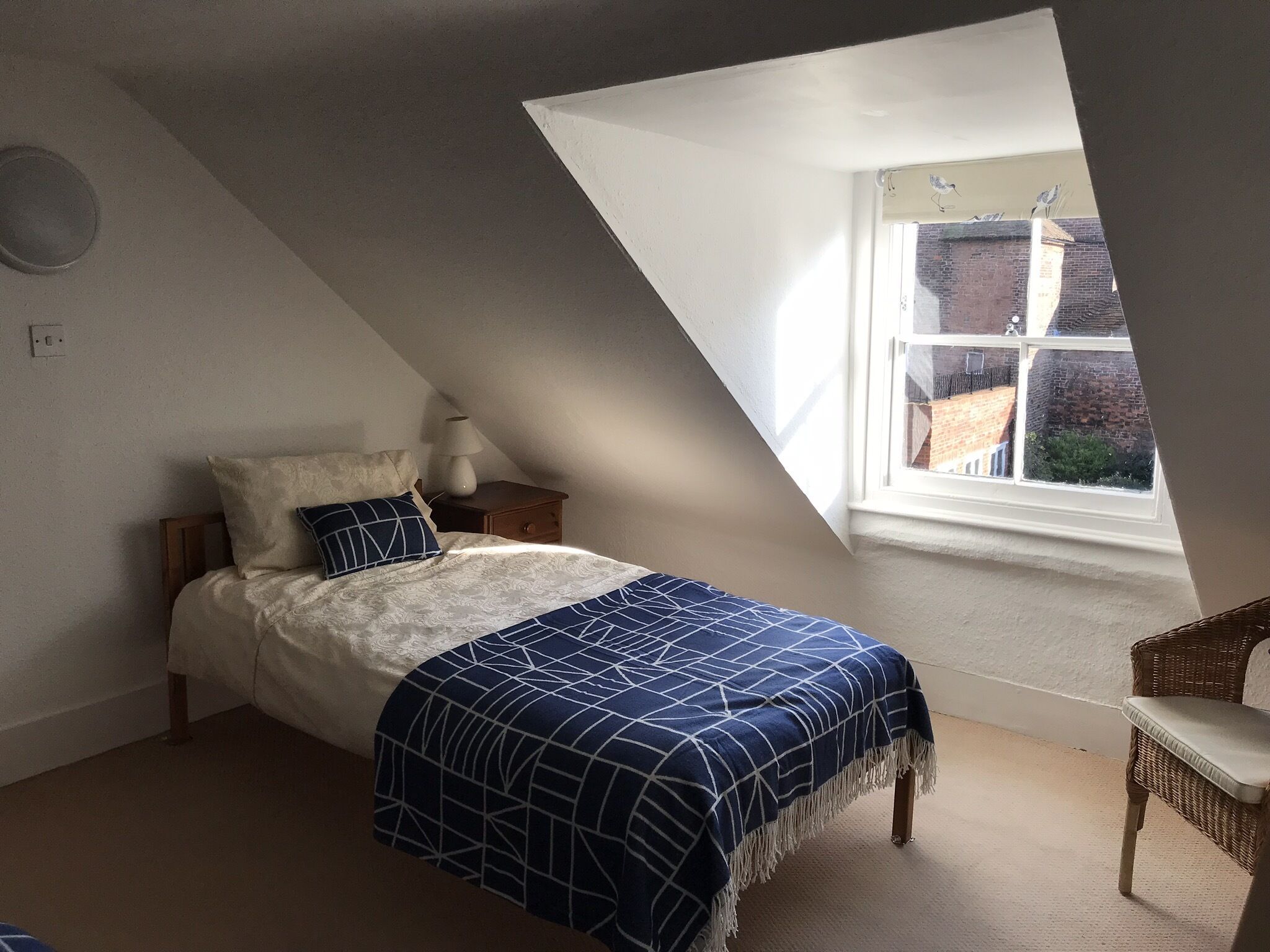 A bedroom with a sloped ceiling, a bed with a blue and white patterned bedspread, a nightstand with a lamp, and a view of a brick building through a window.