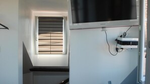 A bedroom with a bed, a TV mounted on the wall, a small bedside table, and a window with blinds.
