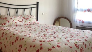 Una cama bien hecha con colcha floral, una pared lisa con dos láminas botánicas enmarcadas, y una ventana con cortinas traslúcidas.
