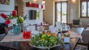 Una mesa de comedor con una fuente de frutas, un jarrón de flores, un tarro de miel y un vaso de bebida roja.