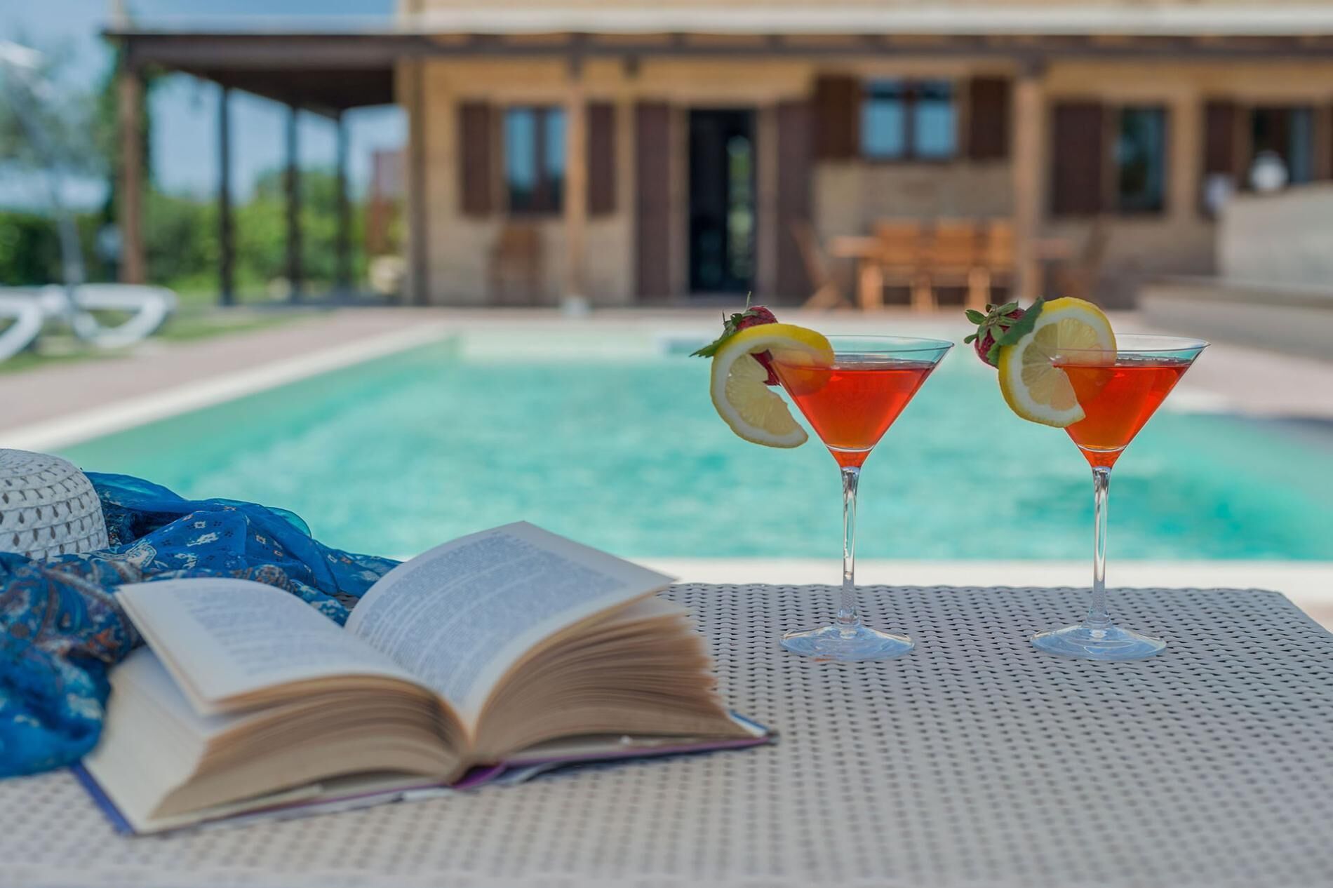 Två martiniglas med frukter som garnering står på ett bord vid poolkanten, en öppen bok ligger bredvid, och i bakgrunden syns en pool med ett hus.
