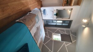 A bedroom with a wooden floor, a bed with patterned bedding, a small table with a mirror, and a window with a view of greenery.