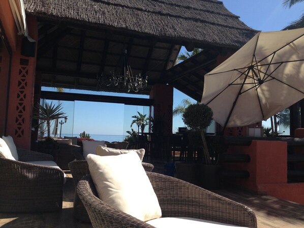 An outdoor seating area with wicker furniture and a large umbrella, overlooking a beach.