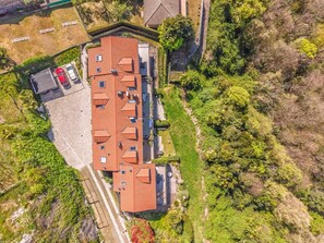 Vista aérea de um edifício com telhado de telhas vermelhas, rodeado por vegetação e uma área pavimentada com um carro vermelho.