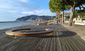 A wooden boardwalk with a circular seating area, trees, and a view of the sea and distant buildings.