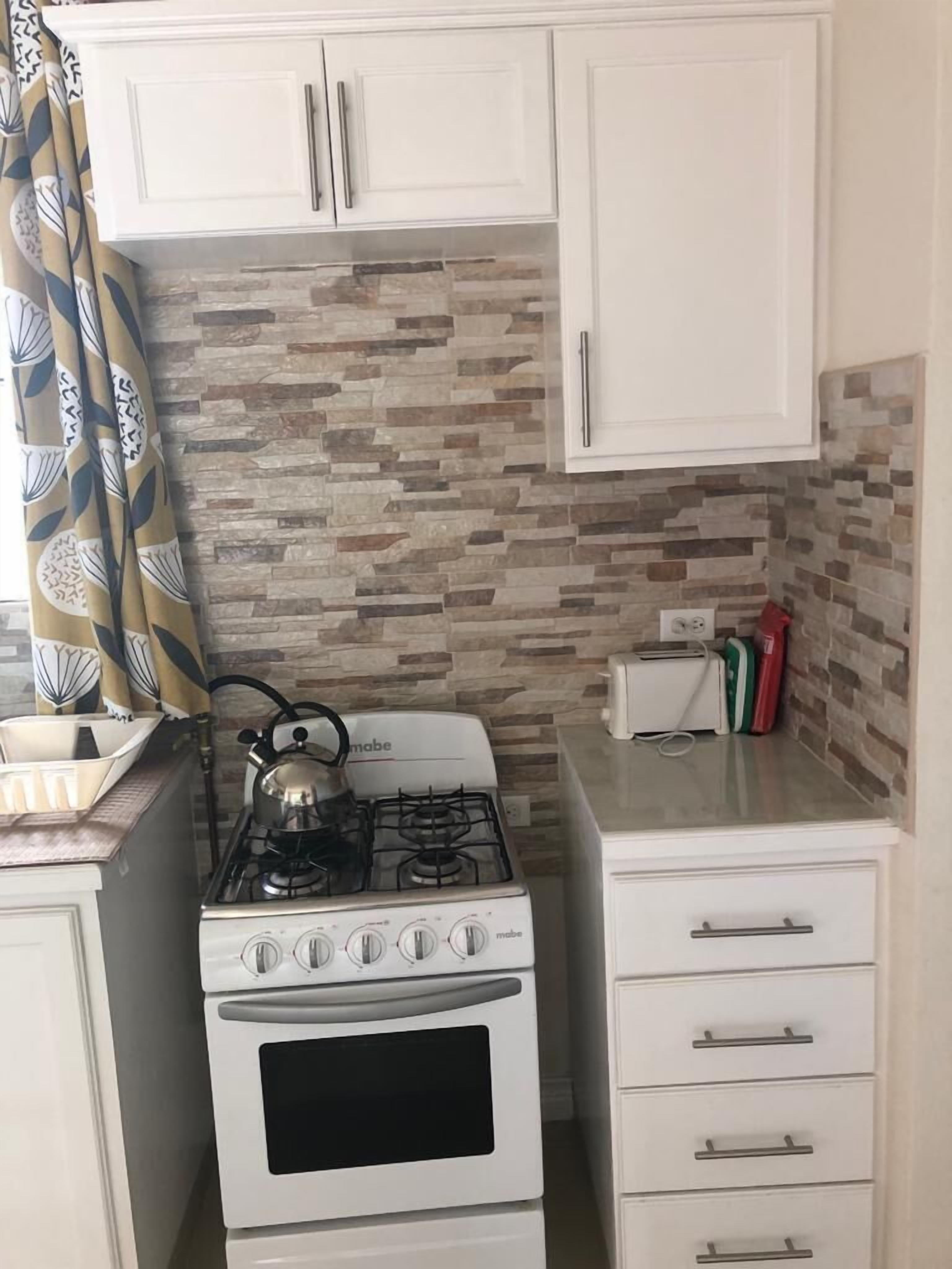 Una cocina compacta con estufa y horno blancos, además de una pequeña encimera. Se observan armarios blancos con tiradores y una cortina con estampado.