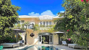 A pool area with lounge chairs and umbrellas, surrounded by lush greenery and a building with a tiled roof.