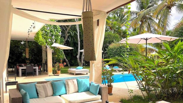 A poolside lounge area with a sofa set, a coffee table with a lantern, and a view of a swimming pool surrounded by lush greenery.
