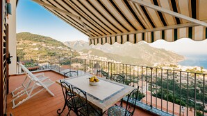 A balcony with a table and chairs, overlooking a scenic landscape with mountains and a coastal view.
