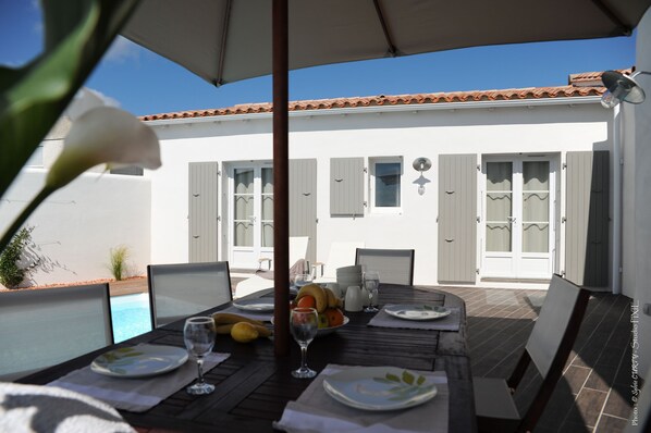 An outdoor dining area with a table set for two, a pool, and a white building with shutters in the background.
