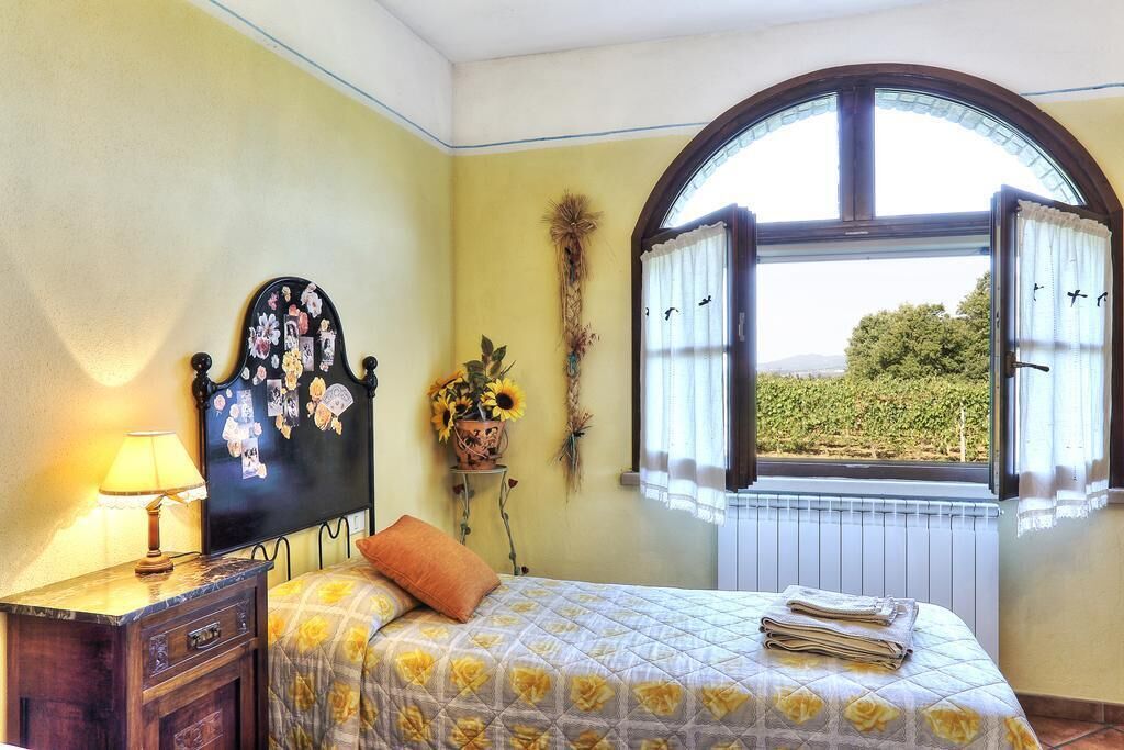 Een slaapkamer met een bed, nachtkastje, lamp, raam met gordijnen en uitzicht op groen.