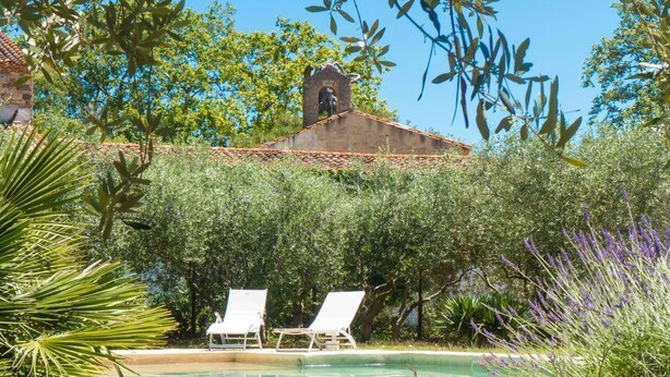 A pool area with two lounge chairs, surrounded by lush greenery and a building with a tiled roof in the background.