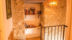A narrow stone-walled corridor with wooden shelves holding books and a hanging lamp.