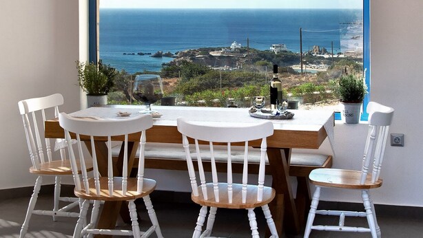 A dining area with a large window overlooking the sea, a wooden table, white chairs, and a hanging light fixture. - AELIA LUXURY VILLA IN A PRIVATE ESTATE IN ARDANI KARPATHOS (Amoopi)