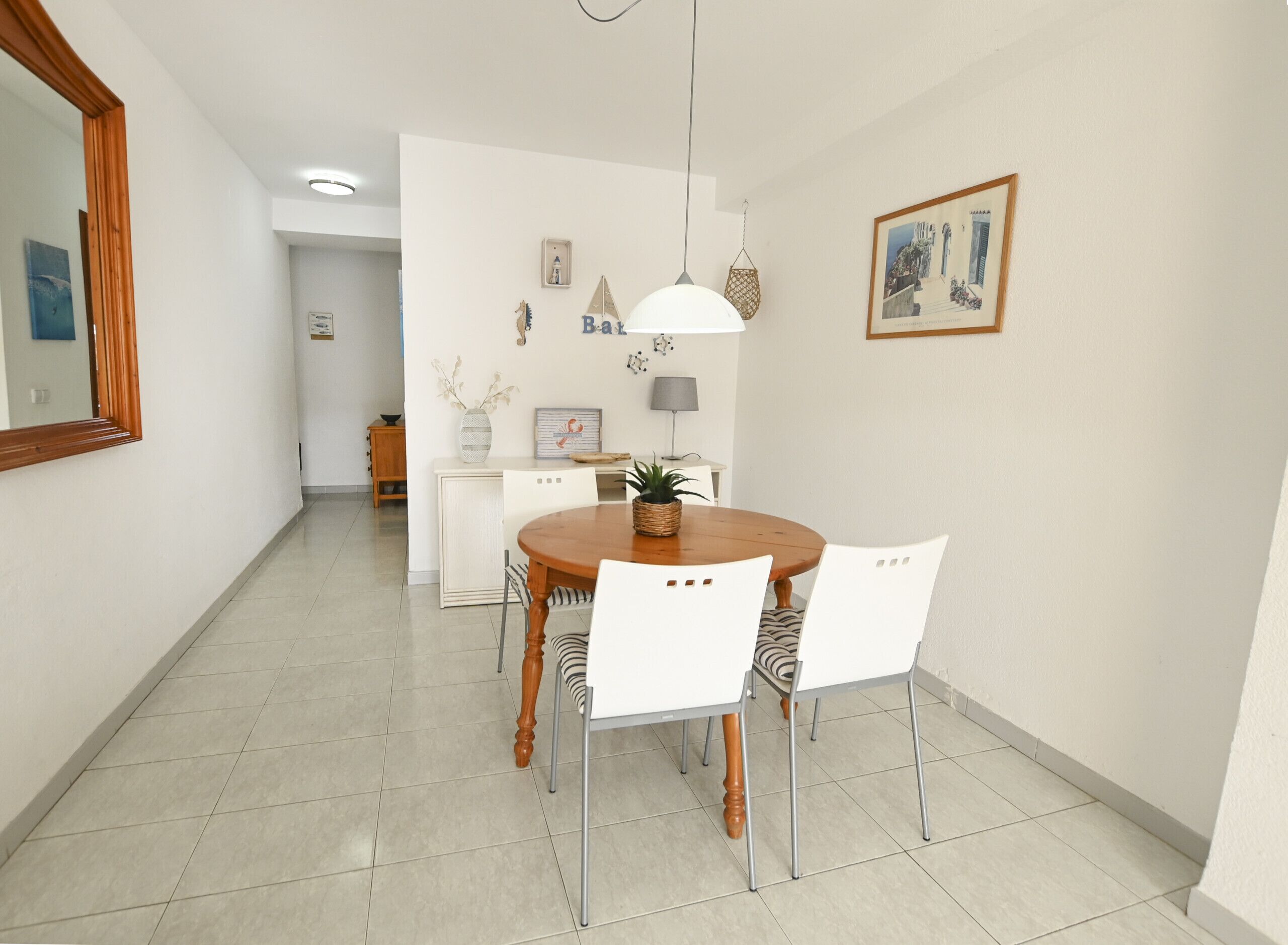 A dining area with a round wooden table, white chairs, a hanging pendant light, and a framed picture on the wall.