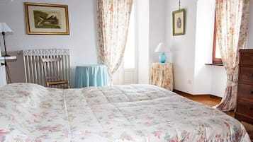 A bedroom with a floral bedspread, a wooden dresser, a chair, and a table with a lamp.