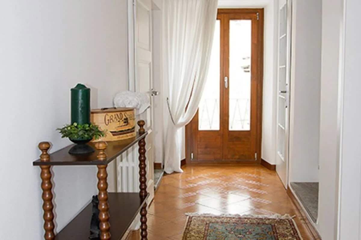 A hallway with a wooden door, white curtains, a small table with a plant, and a patterned rug.