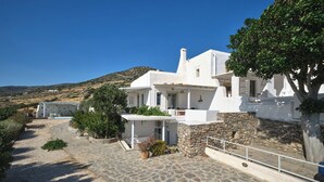 A white building with a stone pathway leading to a door, surrounded by greenery and a clear blue sky. - Modern Cycladic house with pool (Paros)