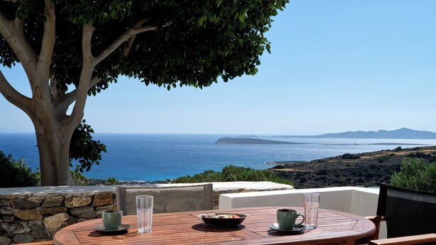 A round wooden table with chairs, a stone wall, and a view of the sea and distant hills. - Modern Cycladic house with pool (Paros)