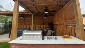 Une cuisine extérieure équipée d’un barbecue, d’un évier et d’un espace détente aménagé sous un toit en bois.