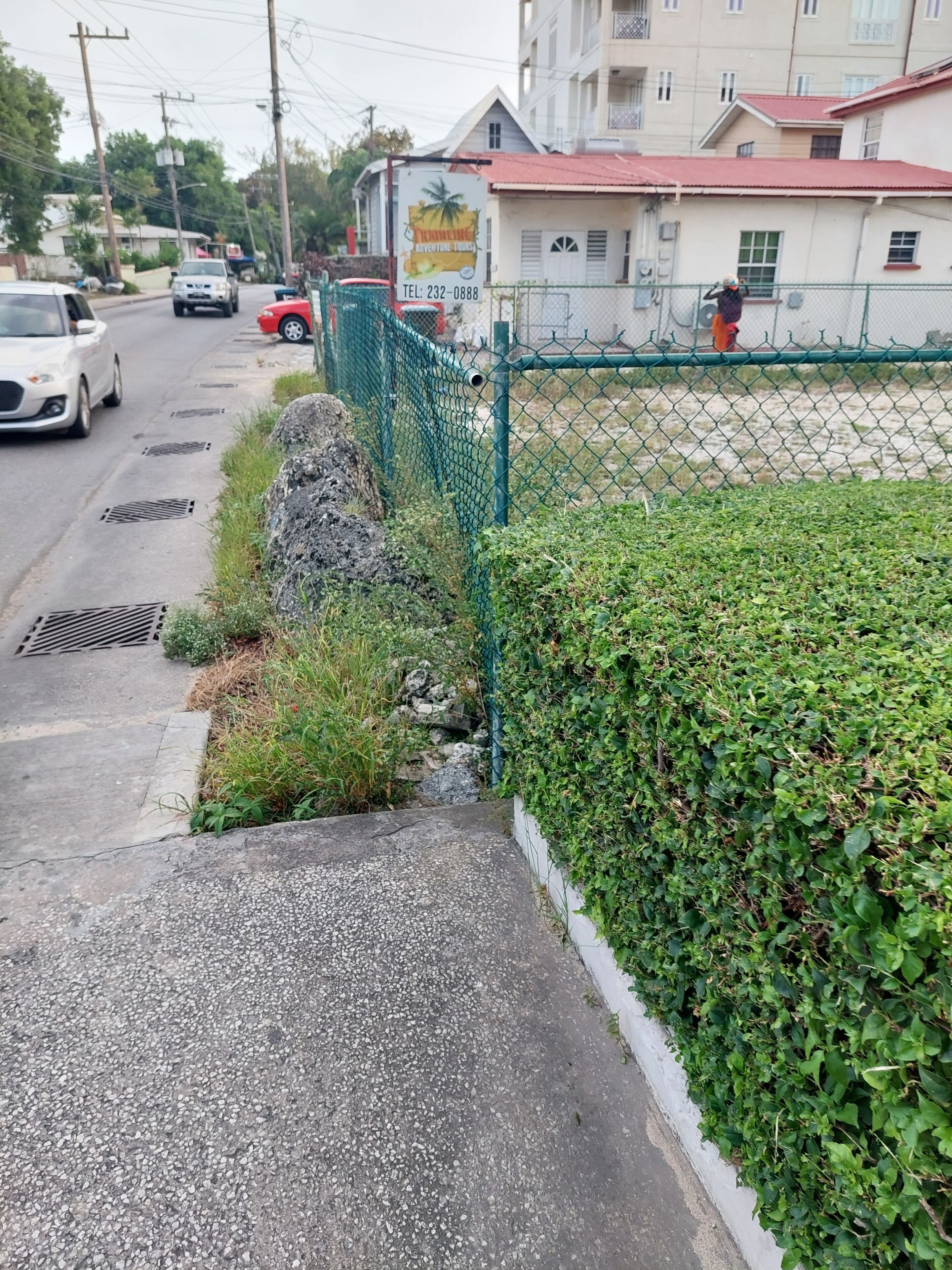A street with a green fence, a white building with a red roof, and a signboard.