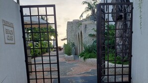 Une entrée fermée par un portail, donnant accès à un club de plage privé, avec un mur en pierre et des plantes tropicales.