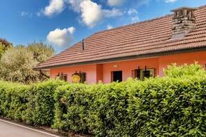 A building with a red-tiled roof, a pink facade, and a well-maintained hedge in front. - Italian Lakes lakeside villa with direct lake access (Ranco)
