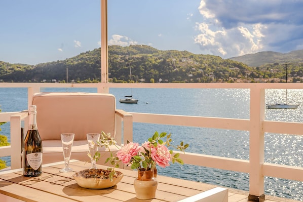 A table with a bottle, glasses, and flowers on a balcony overlooking a body of water with boats and hills in the background.