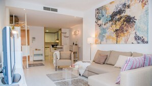 Un salon avec un canapé, une table basse et un téléviseur. Il y a un tableau au mur et une cuisine à l’arrière-plan.