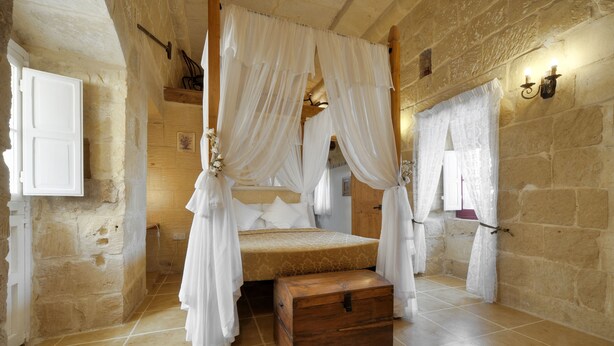 A stone-walled bedroom with a four-poster bed, a wooden chest, and a small window with curtains. - Tal-barun Farmhouse (San Lawrenz)