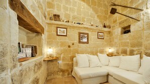 A stone-walled room with a white sofa, shelves with books and decorative items, and a small table with a lamp. - Tal-barun Farmhouse (San Lawrenz)